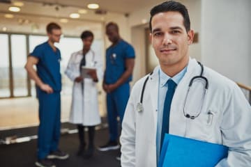 a male doctor in a white coat standing in front of other doctors discussing a case