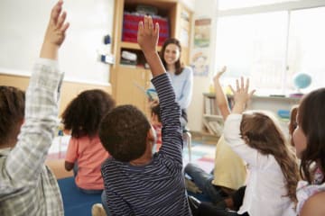 Teacher reads to students sitting on the floor raising their hands