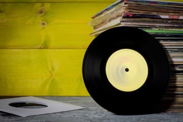 Vinyl record leans against stack of vinyl record sleeves with yellow wood background