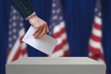 Ballot being placed in voter box with American flags behind the voting station.