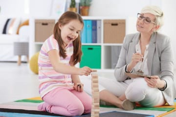 Child counselor sits on floor and observes a little girl playing with wood blocks