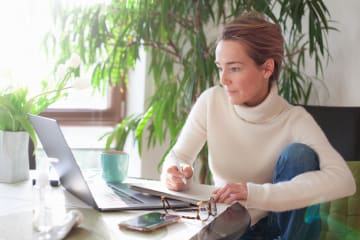 Slender blonde woman takes notes to develop her creative writing story on her laptop