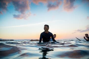 Man smiling while taking in the sunset in the ocean with other surfers