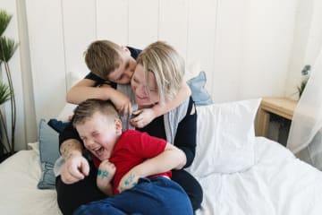 A young son kisses mom on the cheek on her bed and his brother is laughing