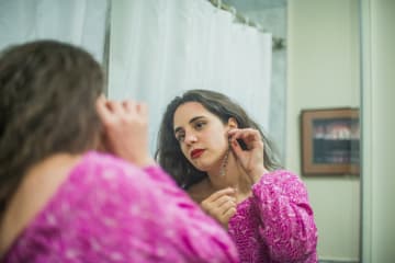 Caucasian brunette woman in pink dress putting earring on while looking in a mirror