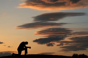 Illumination of a man kneeled on top of a hill praying in front of an ending sunset