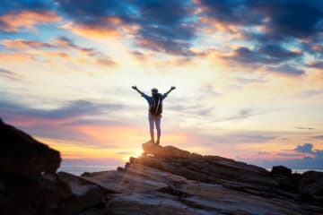 Person stands triumphantly on top of a mountain with sunset in horizon