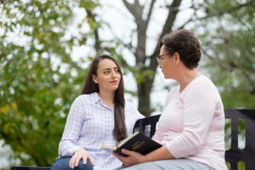 Older woman with Bible open in lap encourages younger woman listening to her