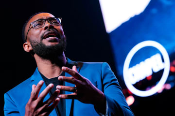 Marcus Doe speaking at GCU chapel - African male in a blue suit with black frame glasses expresses a point at a chapel service