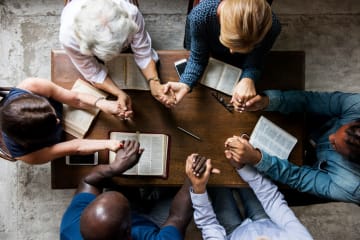 Multicultural group around small table holds hands in prayer