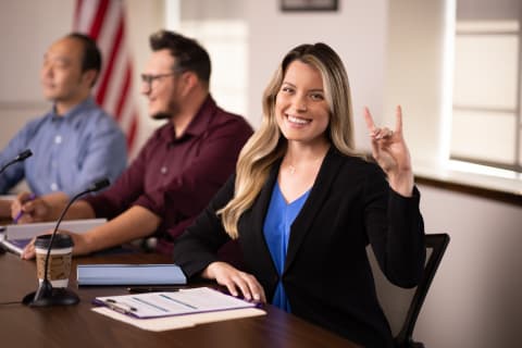 Blonde female public admin student smiling with lopes up sign at table with others