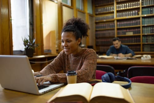 Happy student working in a library with an open book and laptop