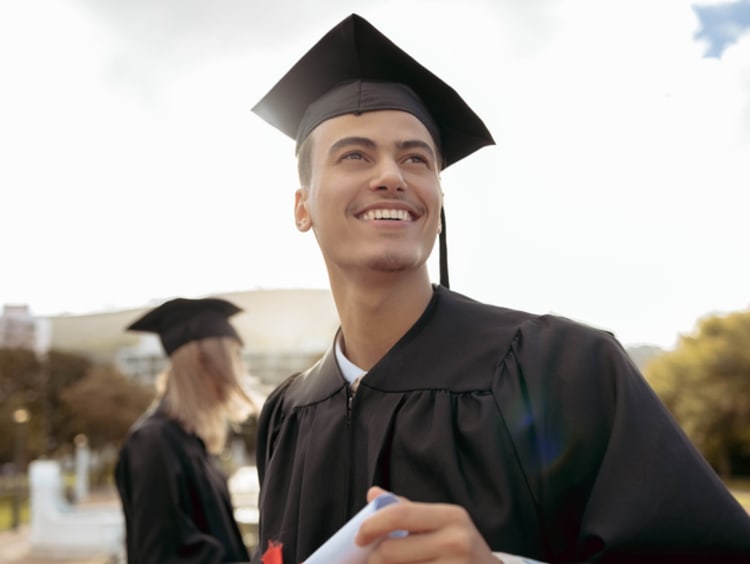 Young man outside dressed in regalia smiling and holding certificate