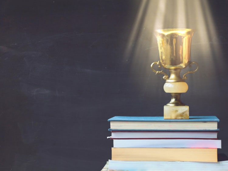 Golden trophy on pile of books, against blackboard, with sun rays over trophy; learning/achievement concept - stock photo