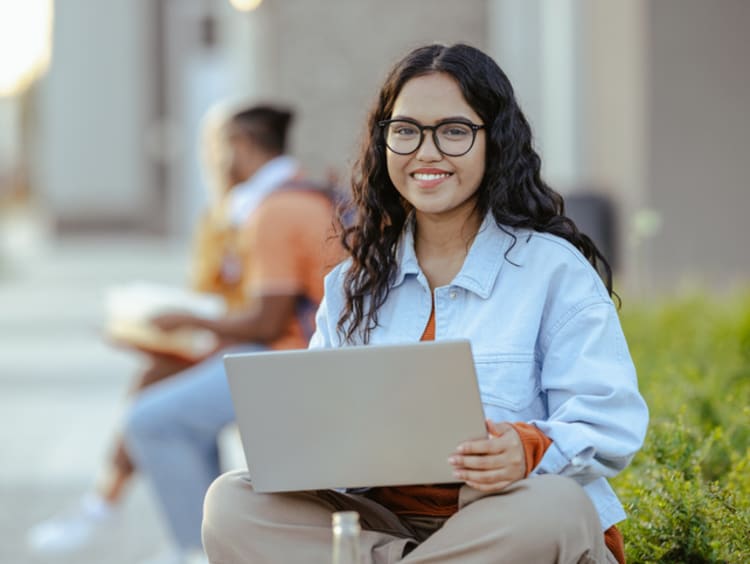Portrait of a young Latin female student using laptop on the bench on campus - stock photo