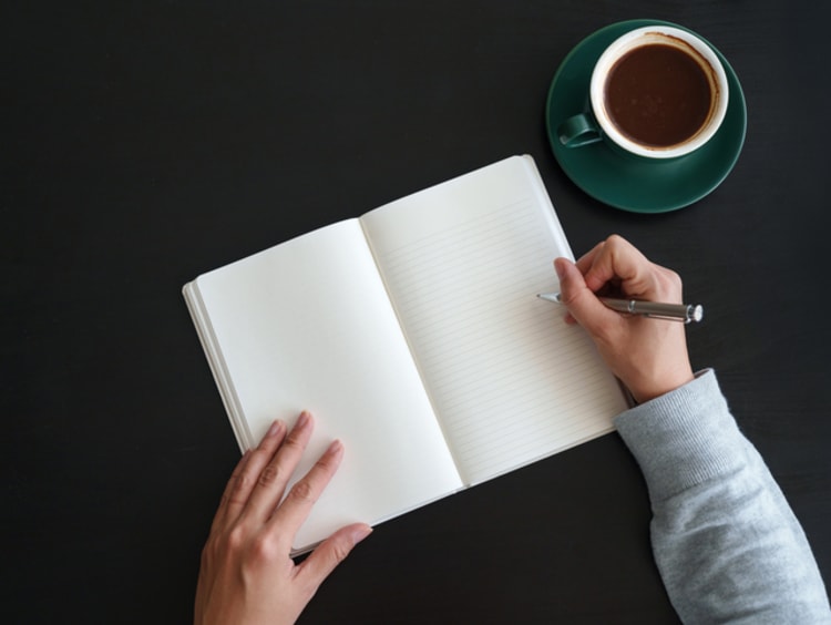 Overhead view of an Asian woman writing in diary while having coffee and working at the desk at home - stock photo