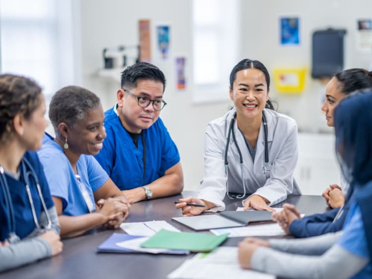 A small group of doctors and nurses gather around a meeting table to discuss patient cases. They are each dressed professionally and have various, files, notes and papers scattered between them as they reference them throughout the meeting.
