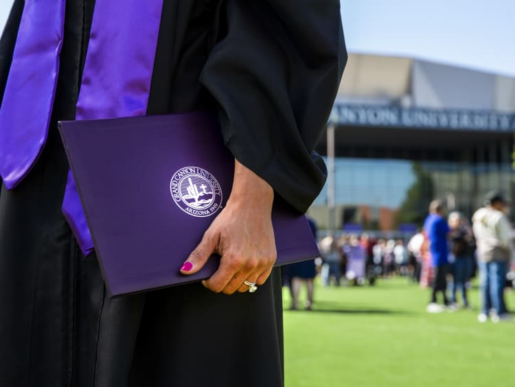 Student in cap and gown holding degree outside the Global Credit Union Arena.