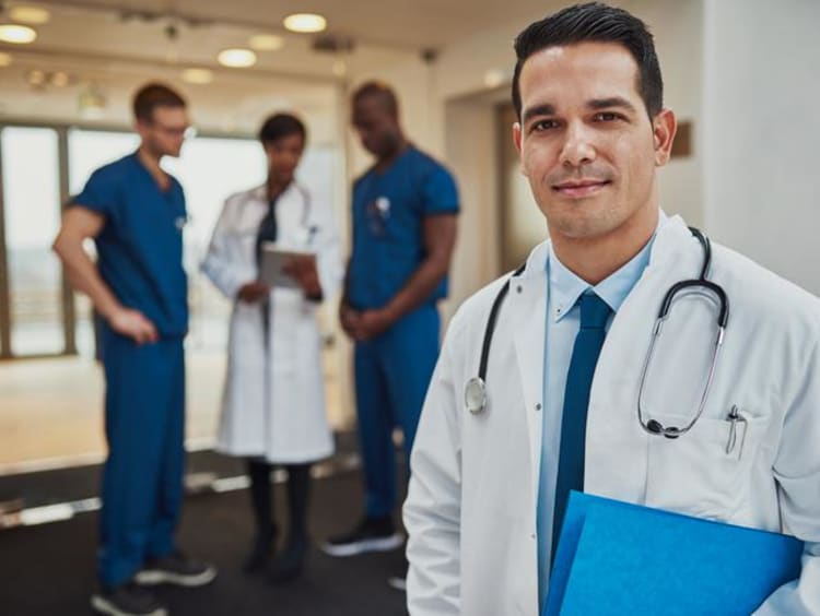 a male doctor in a white coat standing in front of other doctors discussing a case