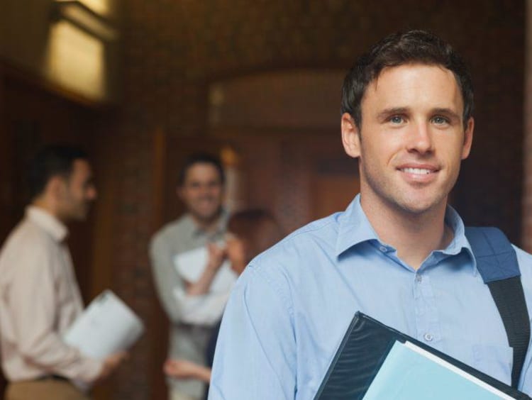 Adult male student holds folder and wears backpack on one shoulder with two males in background