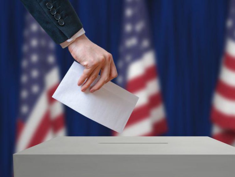 Ballot being placed in voter box with American flags behind the voting station.