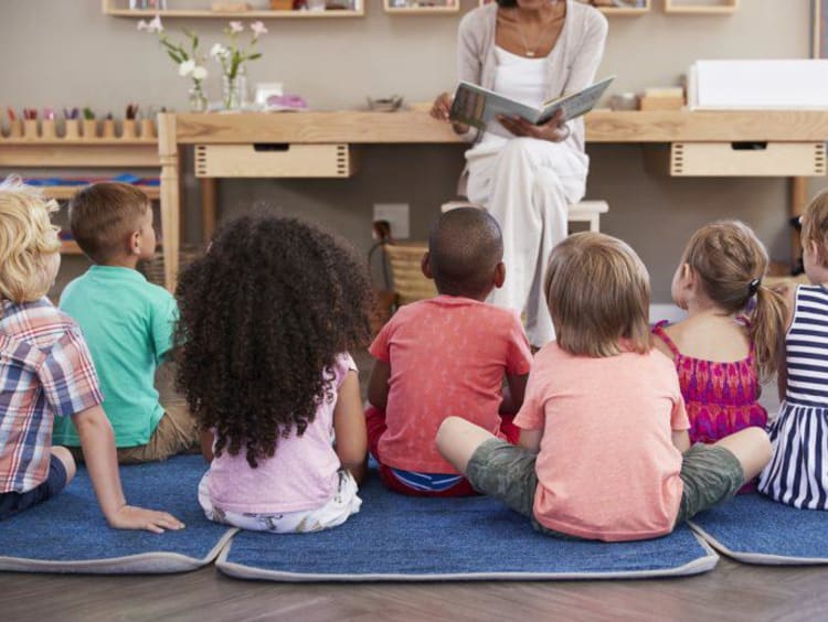 Elementary teacher sits at front of class reading a book to children sitting on ground