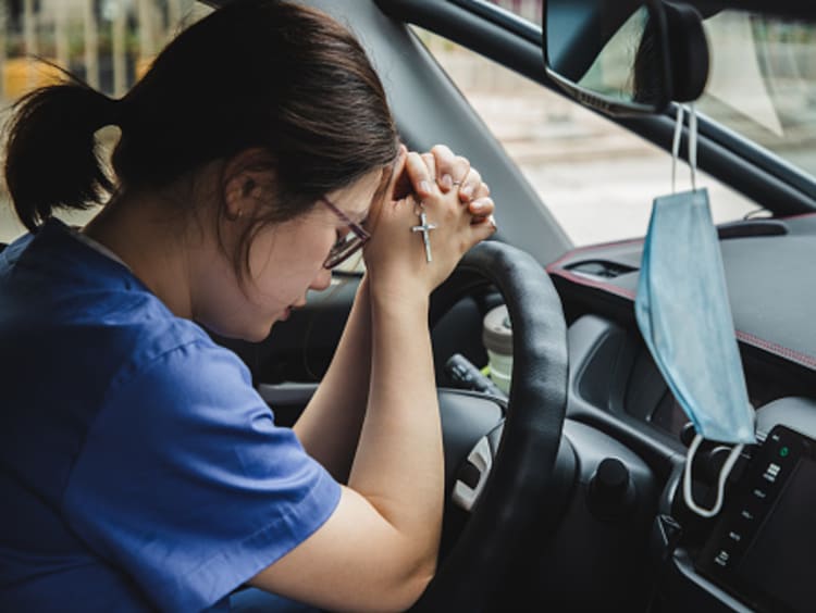 Female brunette nurse in the car praying with mask hanging from rearview mirror