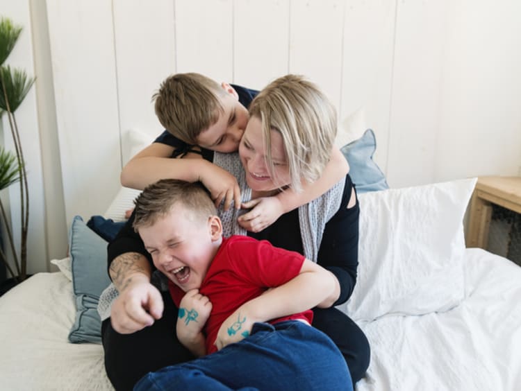 A young son kisses mom on the cheek on her bed and his brother is laughing