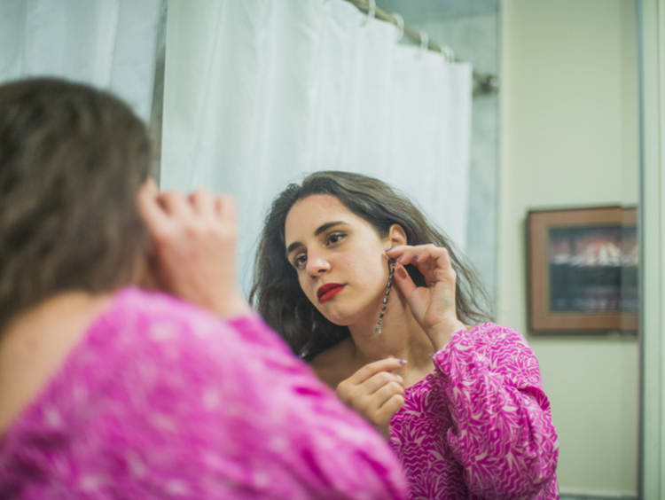 Caucasian brunette woman in pink dress putting earring on while looking in a mirror