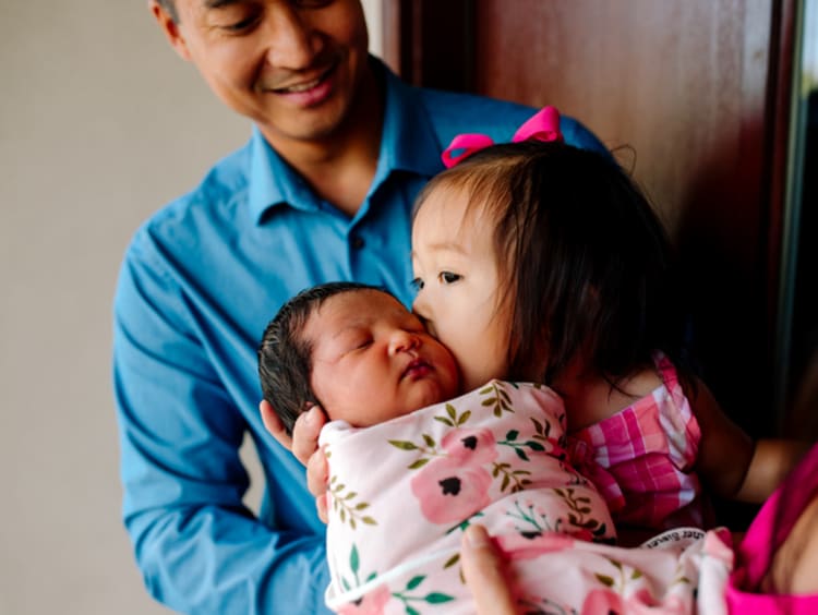 Little Asian girl kissing baby sister on the cheek while man in blue shirt watches