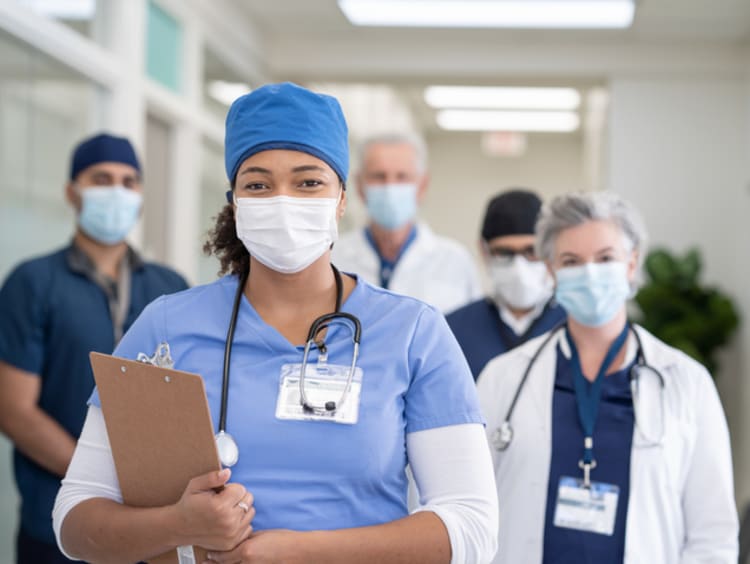 A portrait of a multi ethnic group of healthcare professionals consisting of doctors and nurses at the hospital. A female nurse of African descent is standing in front of the team. Everyone is wearing a face mask and looking at the camera.