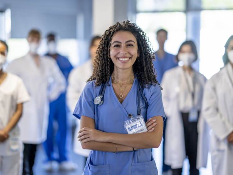 A large group of medical professionals stand in the hallway of a Hospital as they pose together for a portrait. They are each dressed professionally in scrubs and lab coats and some are wearing medical masks to protect them from airborne viruses.