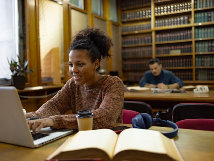 Happy student working in a library with an open book and laptop