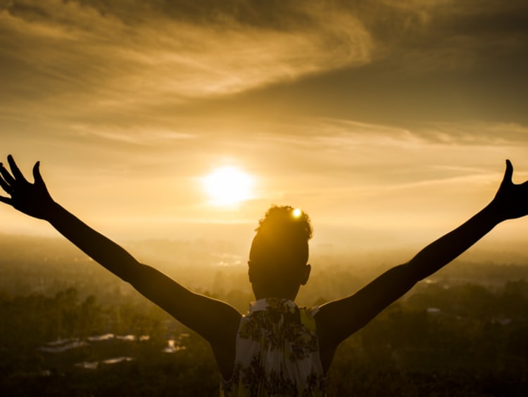 A young African American Woman raises her arms facing the sunset down over the valley and the ocean.