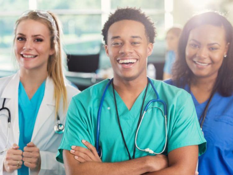 Ethnic nurse male with stethoscope around his neck smiles backed by an African-American female nurse in darker scrubs and a young blonde female in a white lab coat behind his left