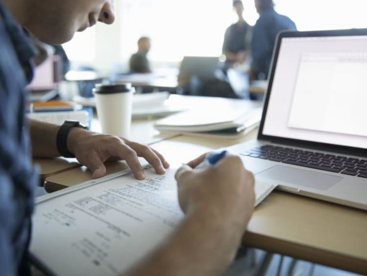 guy writing down in a notebook in front of a laptop