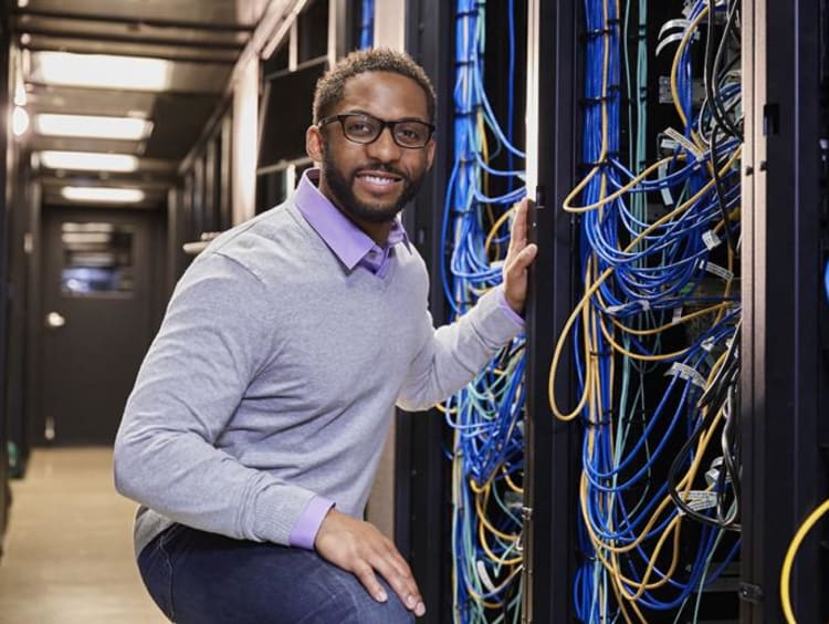 Male African-american IT worker with glasses in light purple sweater kneeling near a server tower