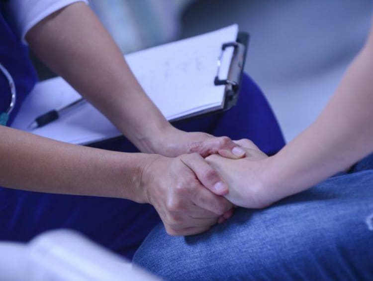 Nurse has clipboard on lap and holds hand of patient