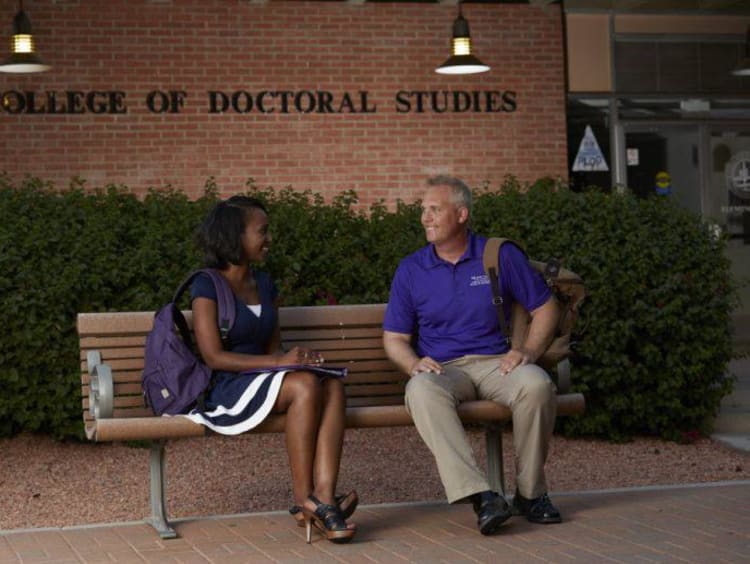 Two students sit on a bench outside the College of Doctoral Studies