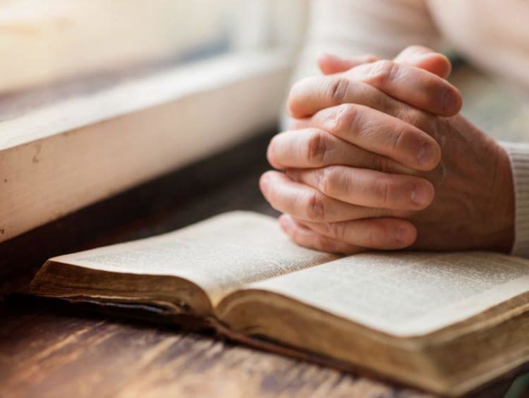 Hands clasped in praying position over open bible next to windowsill