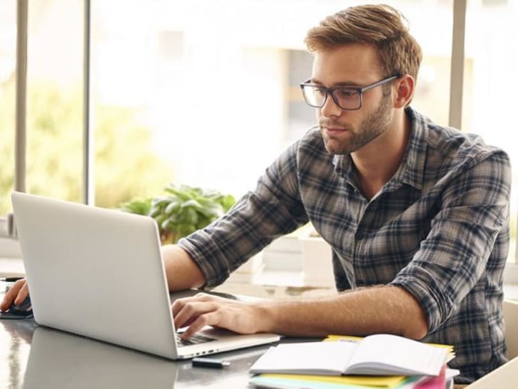 Young male sits at a table working on laptop with coffee and notebooks nearby