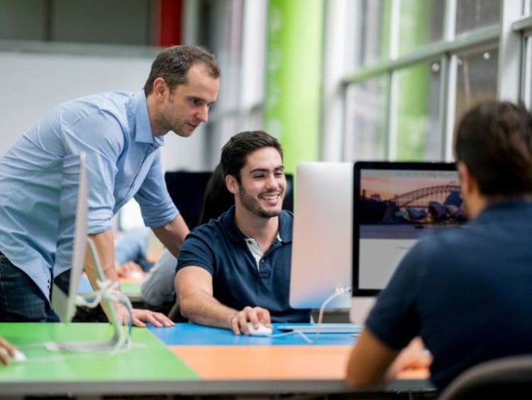 Man sitting down shows guy standing something on his computer monitor