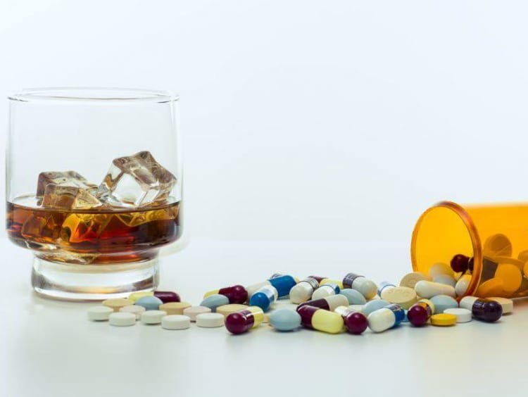 Different pills spill out of bottle next to an alcoholic beverage on ice