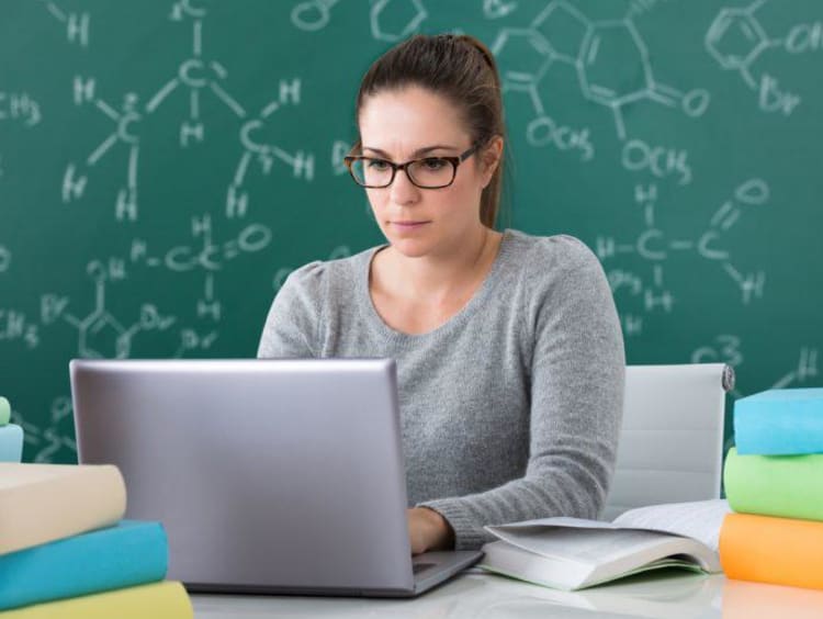 Woman wearing glasses works on laptop with chemistry equations on chalkboard behind her