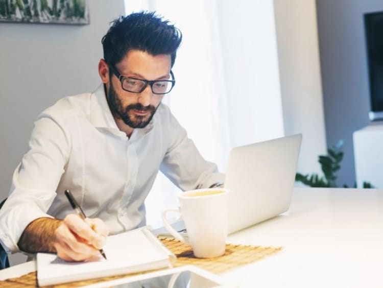 man writing on notebook with laptop in front of him