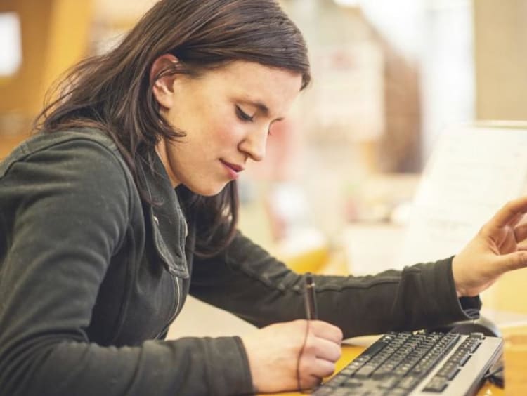 Woman takes notes while pointing at desktop monitor with other hand