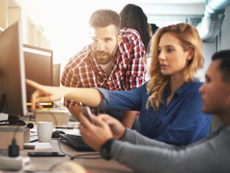 Blonde girl points at computer screen with two others helping her