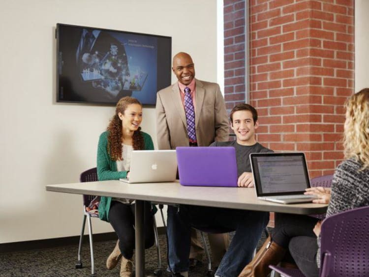 A professor stands behind three students seated and using laptops