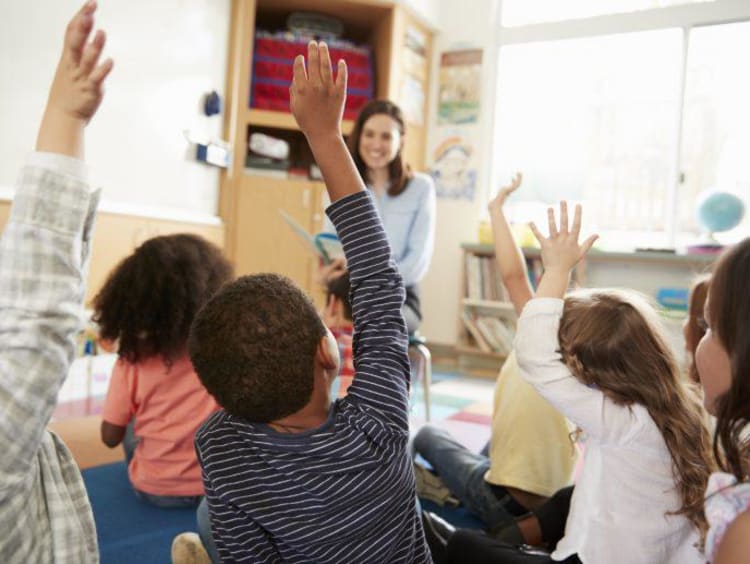 Teacher reads to students sitting on the floor raising their hands