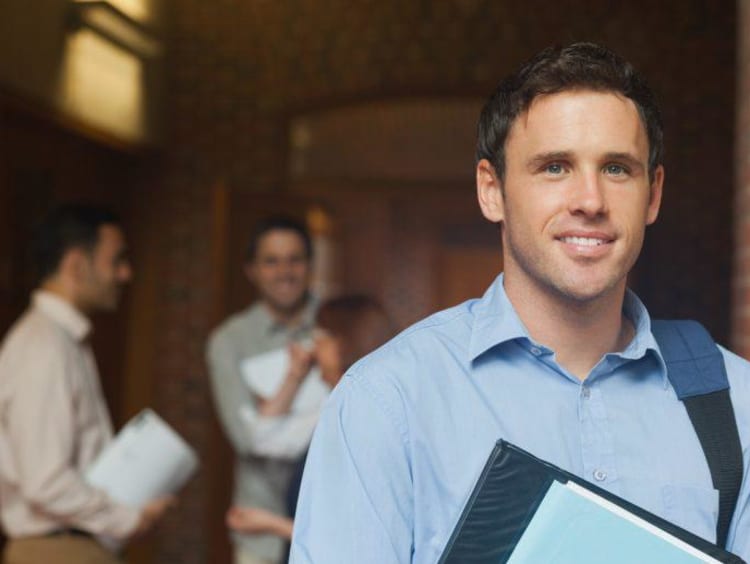 Adult male student holds folder and wears backpack on one shoulder with two males in background